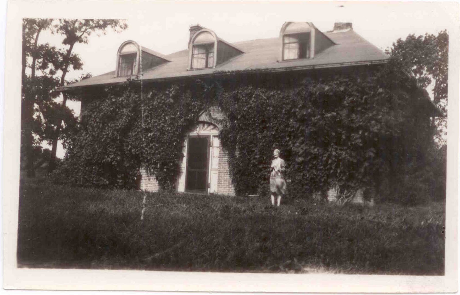 A black-and-white photograph shows a brick house covered in ivy with three dormer windows, and a woman standing in the grassy yard in front of the entrance.