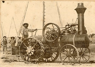 A sepia-toned historical photograph of an early steam-powered traction engine with large spoked wheels and a tall smokestack. A man in a hat and work clothes stands on the engine, while several other workers stand nearby. In the background, wooden oil derricks are visible.