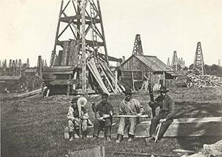 Historical black and white photograph of an early oil field, showing wooden derricks in the background and a group of workers gathered around what appears to be a table or work surface in the foreground. Multiple oil derricks dot the landscape.