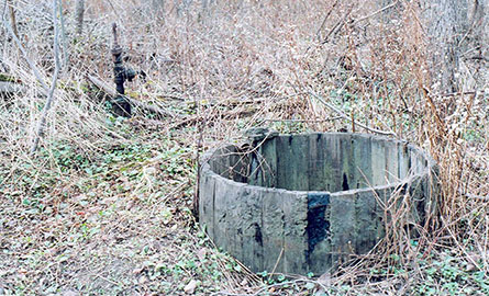 Contemporary color photograph of an abandoned circular well in a wooded area. The well is lined with concrete or stone, and is surrounded by winter vegetation and bare branches. The structure appears weathered and overgrown.