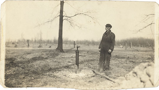 Historical photograph of a man, identified as Frank Wicks, standing beside Gas and Oil Well No. 2 in Oil Springs, circa early 1900s. He is dressed in a dark work outfit and cap, gazing at the well. The landscape is mostly barren with scattered trees and fencing, suggesting a rural oil field setting.