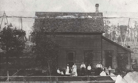 Black and white historical photograph of a wooden house or building with several people standing on the front porch. The building appears to be in an oil field setting, with derricks visible in the background. The structure has a peaked roof with a chimney.
